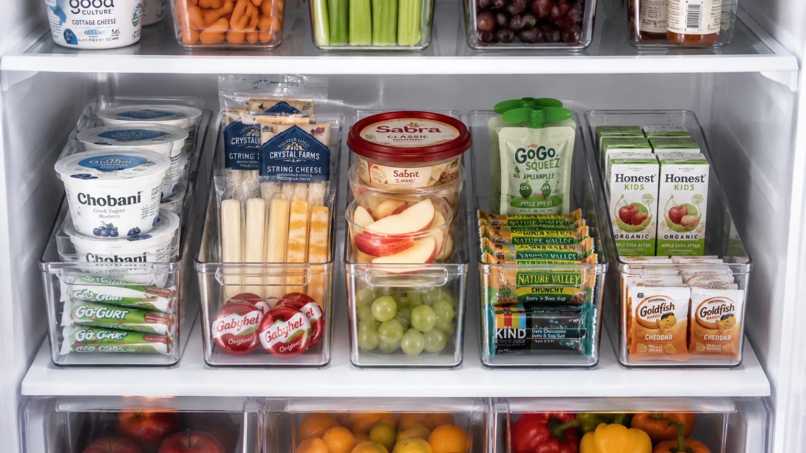 Dedicated refrigerator snack zone with clear bins filled with healthy grab-and-go snacks including yogurt, cheese sticks, fruit, hummus, and juice boxes